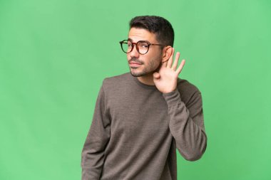 Genç yakışıklı beyaz adam izole edilmiş arka planda bir şeyler dinlerken elini kulağına koydu.