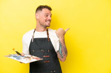 Sarı arka planda izole edilmiş bir palet tutan genç beyaz adam bir ürünü sunmak için yan tarafı işaret ediyor.