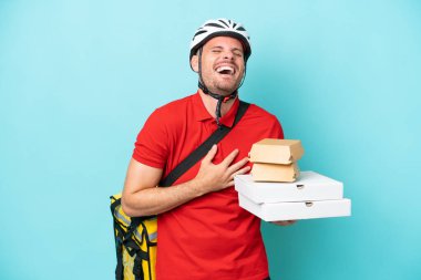 Termal sırt çantalı, fast food taşıyan, mavi arka planda izole edilmiş, sırıtan beyaz bir genç.