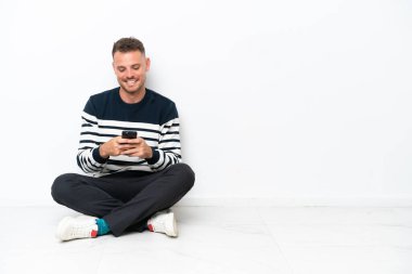 Yerde oturan genç adam beyaz arka planda izole bir şekilde cep telefonuyla bir mesaj gönderiyor.