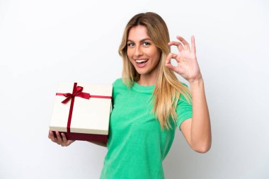 Genç Uruguaylı kadın elinde beyaz arka planda izole edilmiş bir hediye tutuyor. Parmaklarıyla tamam işareti gösteriyor.