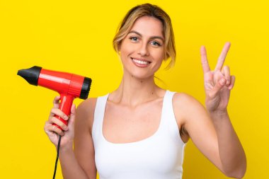 Genç Uruguaylı kadın elinde saç kurutma makinesiyle sarı arka planı izole ediyor gülümsüyor ve zafer işareti gösteriyor.