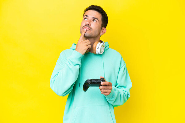 Young handsome man playing with a video game controller over isolated wall having doubts while looking up
