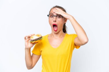 Beyaz arka planda izole edilmiş hamburger tutan genç beyaz bir kadın yan tarafa bakarken sürpriz jest yapıyor.
