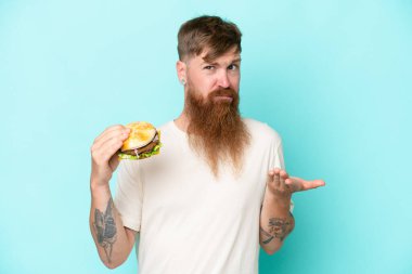 Uzun sakallı kızıl saçlı bir adam mavi arka planda izole edilmiş bir burger tutuyor. Omuzlarını kaldırırken şüphe uyandırıyor.