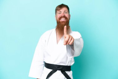 Kızıl saçlı, uzun sakallı, karate yapan, mavi arka planda izole edilmiş, parmağını kaldıran bir adam.