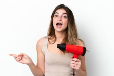 Beyaz arka planda izole edilmiş saç kurutma makinesini tutan beyaz saçlı genç bir kadın şaşırmış ve parmağıyla kenarı işaret ediyor.