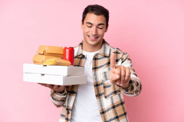 Pembe arka planda fast food tutan genç beyaz adam parmağını kaldırıyor ve gösteriyor.