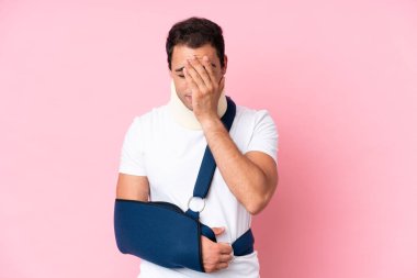 Kırık kollu, pembe arka planda askı takan, yorgun ve hasta bir ifadeyle genç, beyaz bir adam.