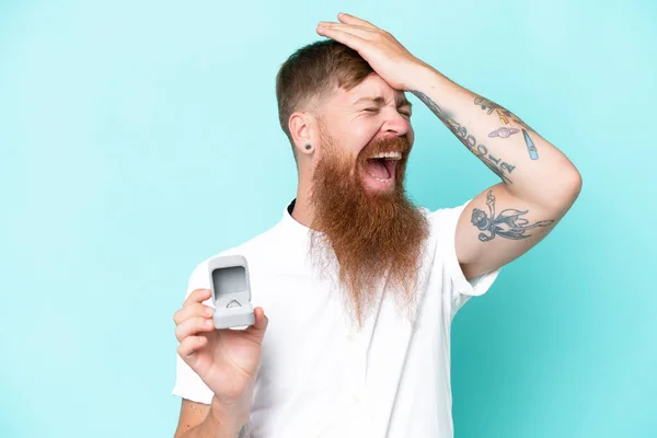 Redhead man with long beard holding a engagement ring isolated on blue background has realized something and intending the solution