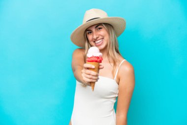 Mavi arka planda izole edilmiş kornet dondurmalı mayolu beyaz kadın mutlu bir ifadeyle.