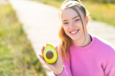 Açık havada elinde mutlu bir ifadeyle avokado tutan genç ve güzel bir kız.