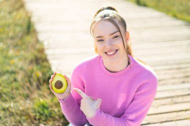 Genç ve güzel bir kız dışarıda bir avokado tutuyor ve onu işaret ediyor.
