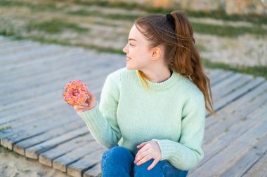 Genç ve güzel bir kız dışarıda elinde mutlu bir ifadeyle bir donut tutuyor.