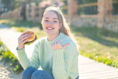 Genç, güzel bir kız elinde hamburgerle bir ürünü sunmak için kenarı işaret ediyor.