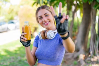 Dışarıda elinde portakal suyu tutan genç ve güzel bir kadın gülümsüyor ve zafer işareti gösteriyor.