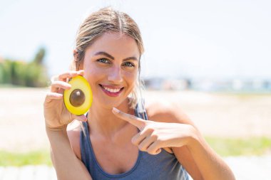 Sarışın genç bir kadın dışarıda bir avokado tutuyor ve onu işaret ediyor.