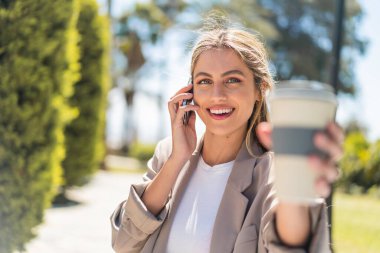 Genç, güzel sarışın Uruguaylı kadın açık havada cep telefonu kullanıyor ve elinde mutlu bir ifadeyle kahve tutuyor.