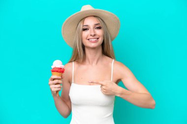 Blonde woman in swimsuit holding an ice cream isolated on blue background and pointing it