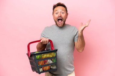 Elinde alışveriş sepeti olan orta yaşlı beyaz bir adam pembe arka planda, şok olmuş bir yüz ifadesiyle izole edilmiş.