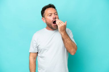 Orta yaşlı, beyaz bir adam mavi arka planda izole edilmiş esniyor ve geniş bir ağzı eliyle kapatıyor.