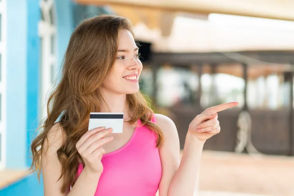 Young redhead woman holding a credit card at outdoors pointing to the side to present a product