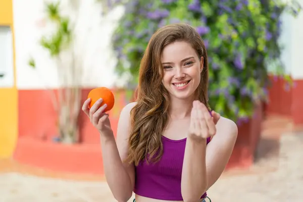 Young redhead woman holding an orange at outdoors inviting to come with hand. Happy that you came