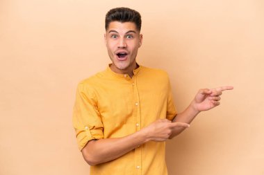 Young caucasian man isolated on beige background surprised and pointing side