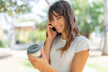 Dışarıda cep telefonu kullanan ve elinde mutlu bir ifadeyle kahve tutan genç bir kadın.