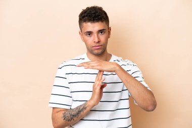 Genç Brezilyalı adam bej arka planda izole edilmiş, zaman aşımı hareketi yapıyor.