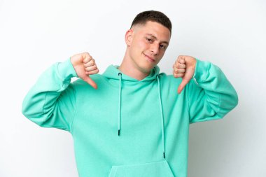 Beyaz arka planda izole edilmiş genç Brezilyalı adam baş parmağını iki eliyle aşağı gösteriyor.