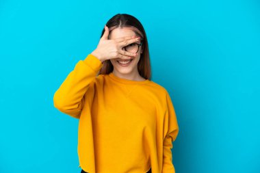 Mavi arka planda izole edilmiş genç beyaz bir kadın. Gözleri elleriyle kaplıyor ve gülümsüyor.