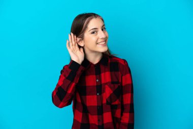 Mavi arka planda izole edilmiş genç beyaz kadın elini kulağına koyarak bir şeyler dinliyor.