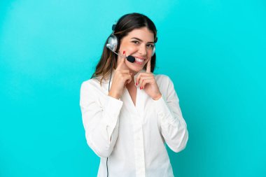 Tele pazarlamacı İtalyan kadın mavi arka planda izole edilmiş kulaklıkla çalışıyor mutlu ve hoş bir ifadeyle gülümsüyor.