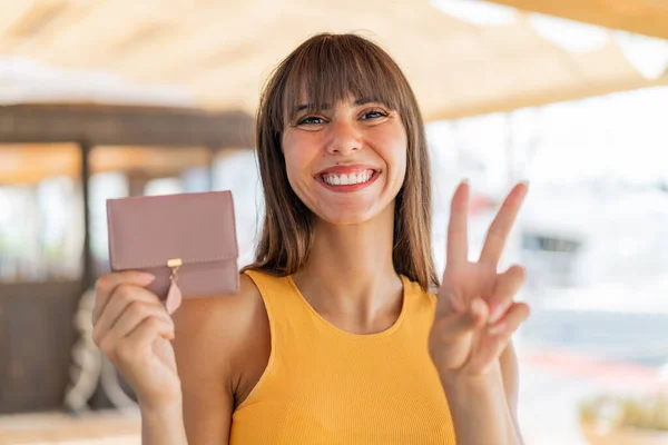 屋外で財布を持っている若い女性は微笑み 勝利の兆候を示しています — ストック写真