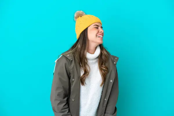 Young Italian woman wearing winter jacket and hat isolated on blue background laughing in lateral position