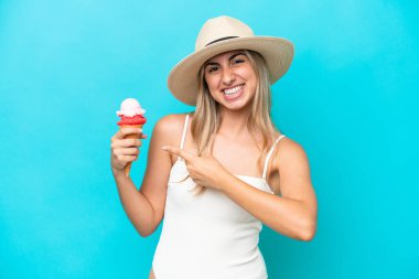 Young caucasian woman in swimsuit with a cornet ice cream isolated on blue background and pointing it