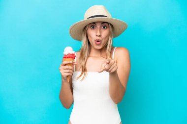 Mavi arka planda izole edilmiş kornet dondurması olan mayo giymiş beyaz bir kadın şaşırmış ve ön tarafı işaret ediyor.