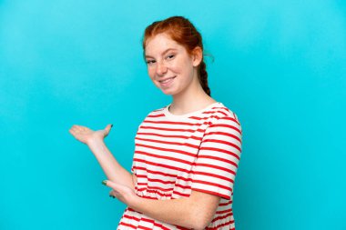 Young reddish woman isolated on blue background extending hands to the side for inviting to come