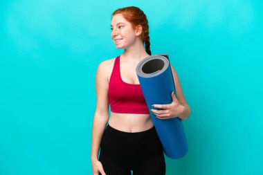 Genç spor kırmızısı kadın yoga derslerine giderken mavi arka planda bir minder tutuyor ve yan tarafa bakıyor ve gülümsüyor.