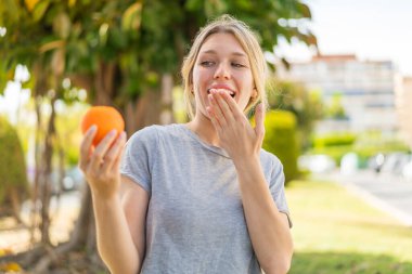 Açık havada elinde portakal tutan genç sarışın kadın. Şaşırmış ve şok olmuş bir yüz ifadesi var.