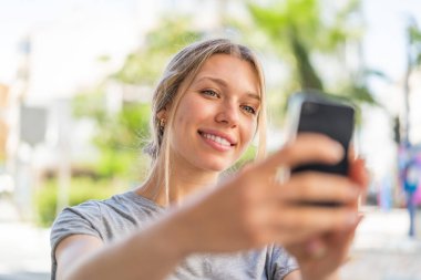 Açık havada genç sarışın bir kadın cep telefonuyla selfie çekiyor.