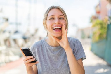 Açık havada cep telefonu kullanan genç sarışın kadın ağzı açık bağırıyor.