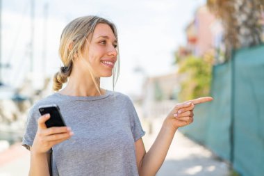 Açık havada cep telefonu kullanan genç sarışın bir kadın bir ürün sunmak için yan tarafı işaret ediyor.