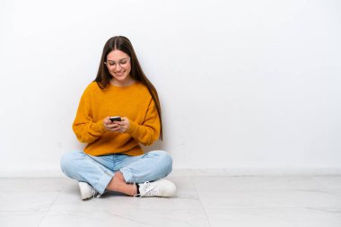 Yerde oturan genç kız beyaz arka planda izole bir şekilde cep telefonuyla bir mesaj gönderiyor.