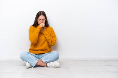 Beyaz arka planda izole bir şekilde yerde oturan genç kız öksürükten muzdarip ve kötü hissediyor.