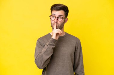 Sarı arka planda izole edilmiş genç beyaz bir adam sessizlik işareti gösteriyor. Parmağını ağzına sokuyor.