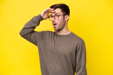 Sarı arka planda izole edilmiş genç beyaz adam yan tarafa bakarken sürpriz jest yapıyor.