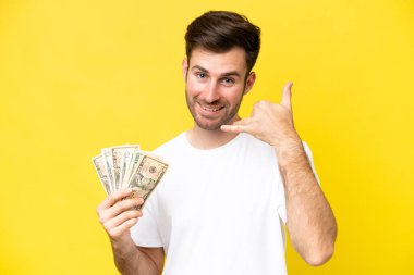 Kafkasyalı genç bir adam telefon hareketleriyle sarı arka planda izole edilmiş bir sürü para alıyor. Bana geri dönüş işareti ver.