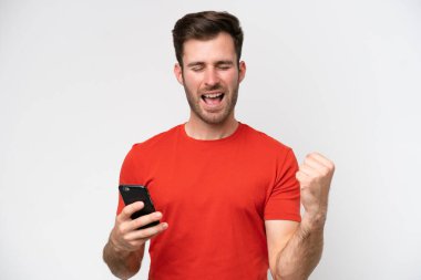 Beyaz arka planda izole edilmiş genç beyaz bir adam cep telefonu kullanıyor ve zafer hareketi yapıyor.